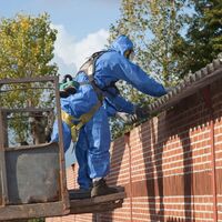 Twee mannen in blauwe overall met een masker op op een hoogwerker die asbest dakplaten vasthouden.