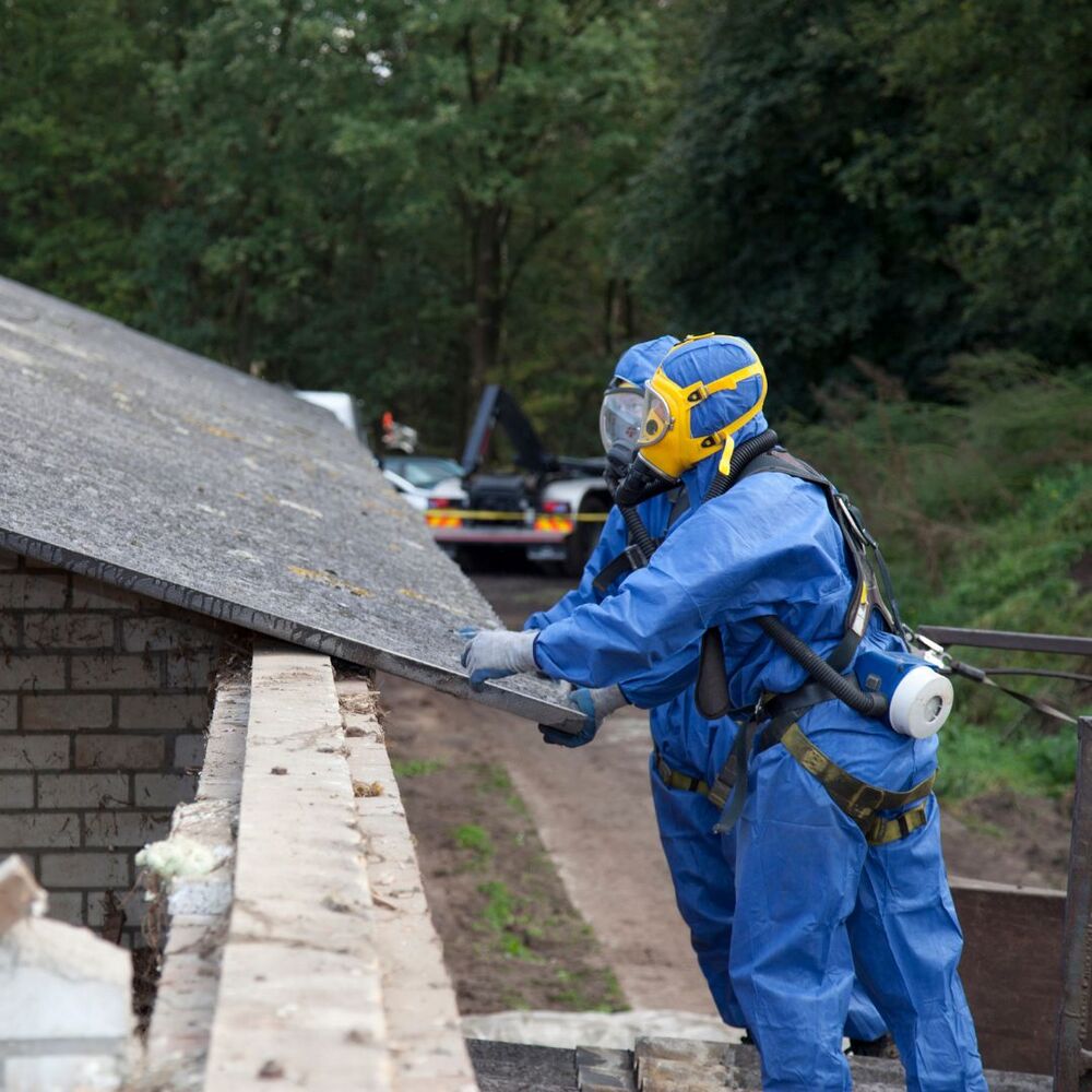 Twee mannen in blauwe overall met een adembeschermingsmasker op. Zij houden dakplaten vast.